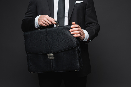 cropped view of businessman in suit holding leather briefcase on dark grey