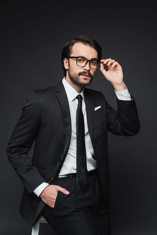 businessman adjusting glasses and leaning on chair while posing with hand in pocket on dark grey