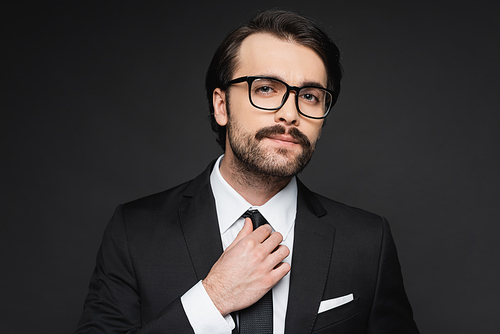 businessman with mustache in glasses adjusting tie on dark grey