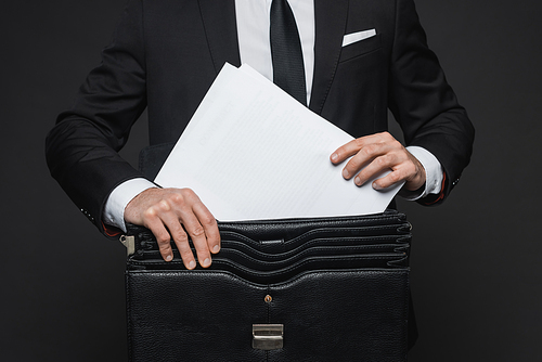 cropped view of businessman putting documents in leather briefcase on dark grey
