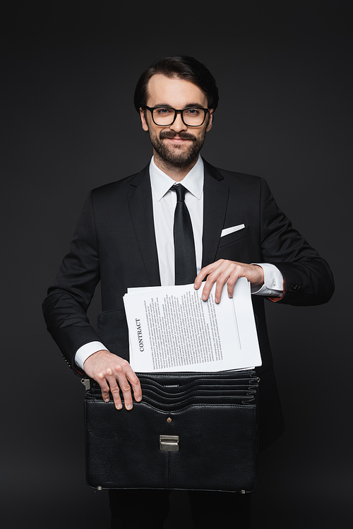 cheerful businessman with mustache in glasses putting contract in leather briefcase on dark grey
