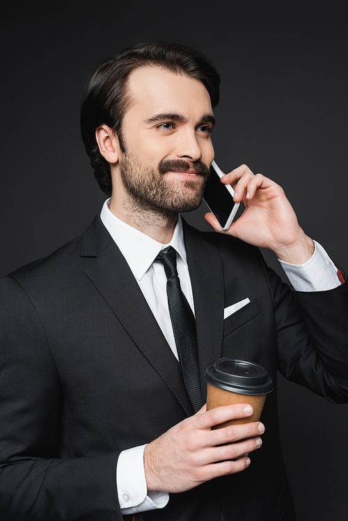 smiling businessman with mustache holding paper cup and talking on smartphone on dark grey