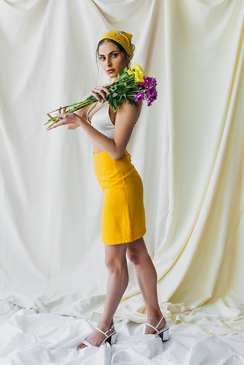 full length of pretty woman in yellow headscarf holding flowers on white