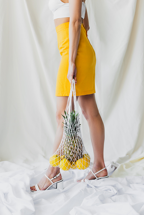 partial view of woman in yellow skirt and crop top holding string bag with lemons and pineapple on white