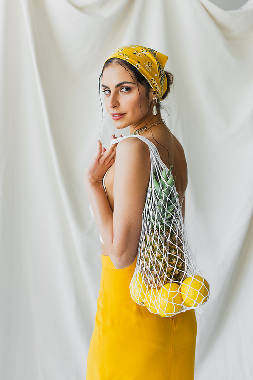 woman in yellow headscarf and crop top holding string bag with lemons and pineapple on white