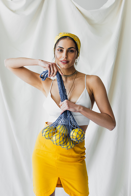 happy woman in yellow headscarf and crop top holding string bag with lemons on white