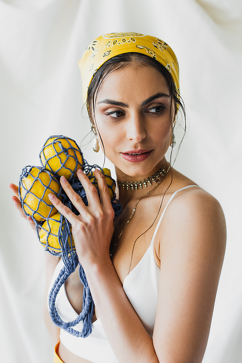 woman in yellow headscarf and crop top holding string bag with lemons on white