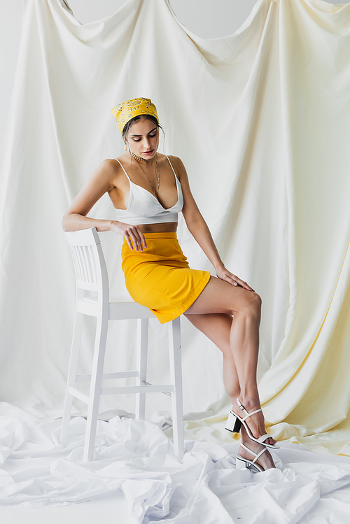 full length of stylish woman in yellow headscarf, skirt and crop top sitting on chair on white