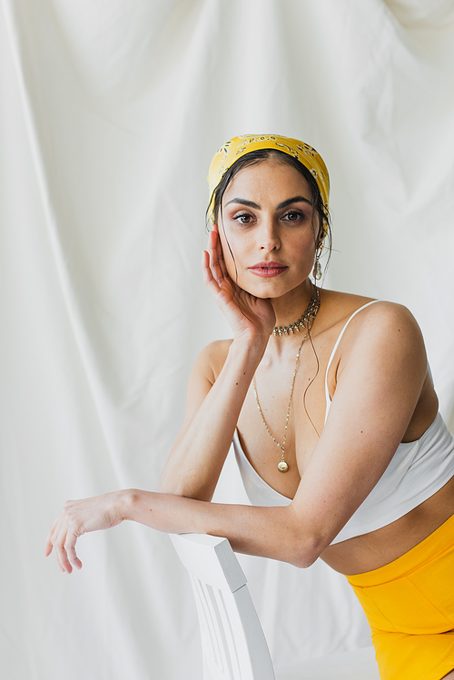 trendy woman in yellow headscarf and crop top sitting on chair on white