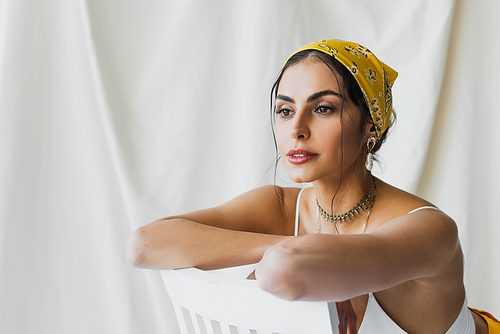 trendy woman in yellow headscarf and crop top leaning on chair on white