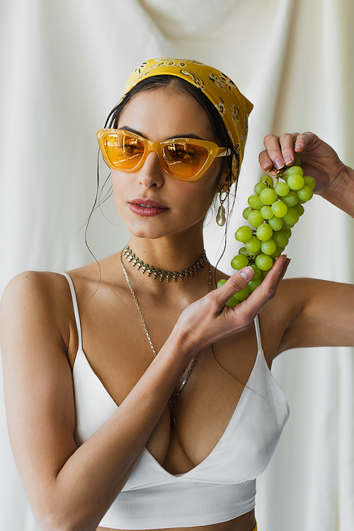 trendy woman in yellow sunglasses, headscarf and crop top holding grapes on white