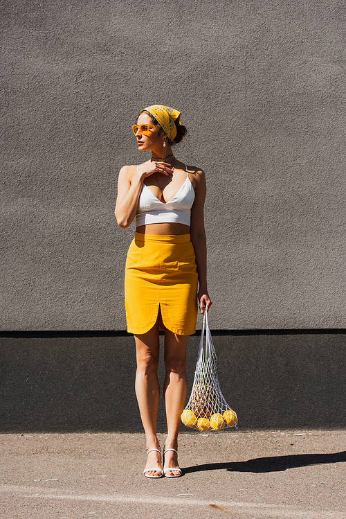 full length of stylish woman in sunglasses and yellow headscarf holding string bag with fruits near concrete wall
