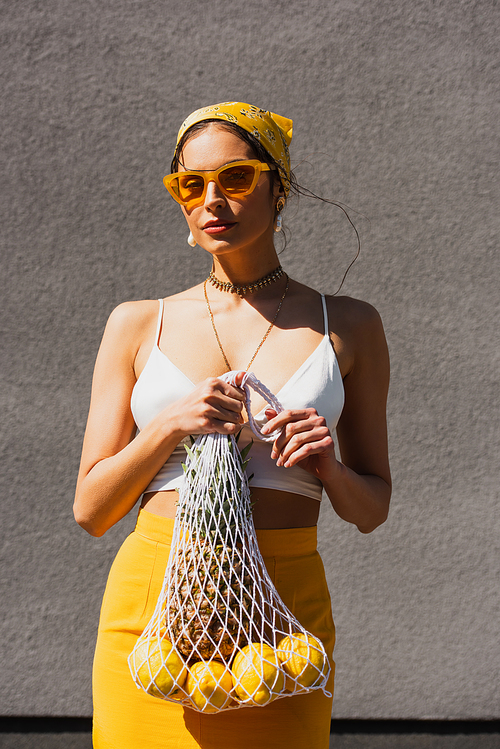 stylish woman in yellow sunglasses and headscarf holding string bag with fresh fruits near concrete wall