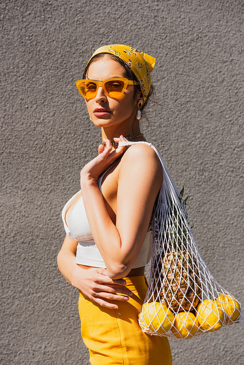 trendy woman in sunglasses and yellow headscarf holding string bag with fruits near concrete wall