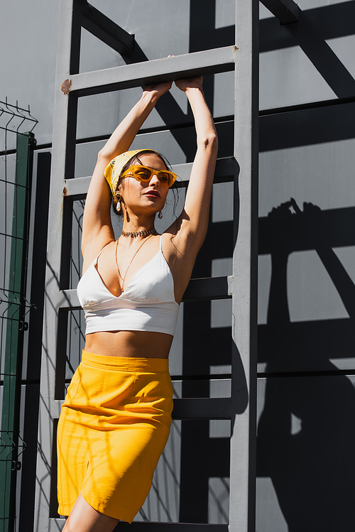 pretty woman in sunglasses and yellow headscarf posing near ladder and concrete wall