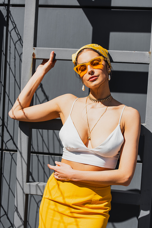 trendy woman in sunglasses and yellow headscarf posing near concrete wall