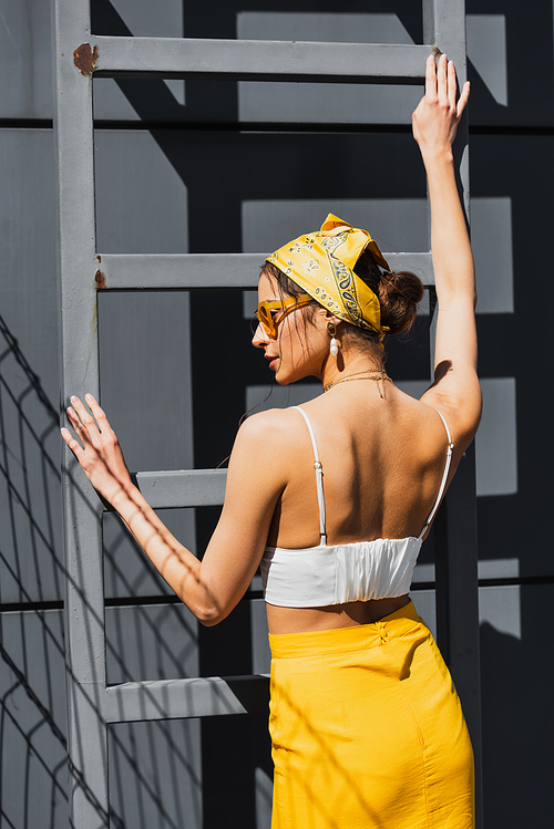 stylish woman in sunglasses and yellow headscarf posing near ladder and concrete wall