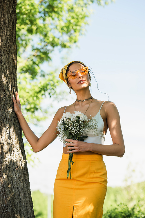 stylish woman in sunglasses and yellow headscarf holding flowers and standing near tree trunk