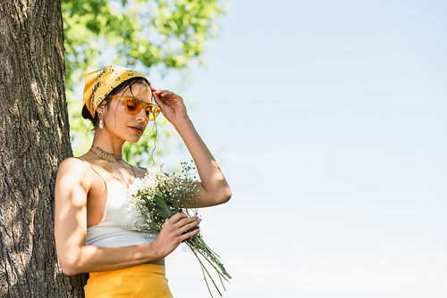 trendy woman in sunglasses and yellow headscarf holding flowers and standing near tree trunk