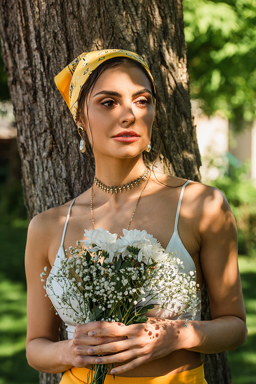 stylish woman in yellow headscarf holding flowers near tree