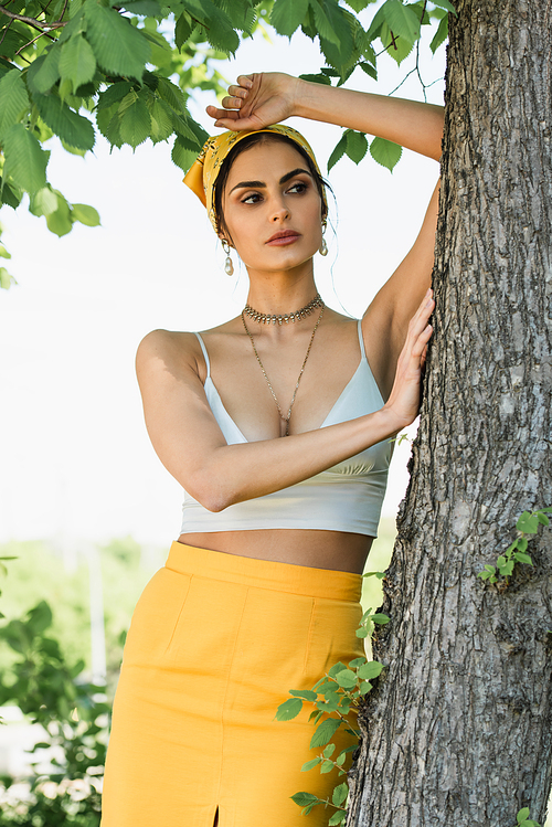 trendy woman in yellow headscarf leaning on tree trunk
