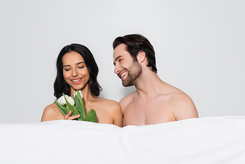 smiling sensual woman holding tulips near shirtless man and white blanket isolated on grey