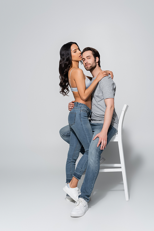 passionate woman in bra and jeans hugging young man sitting on chair and  on grey