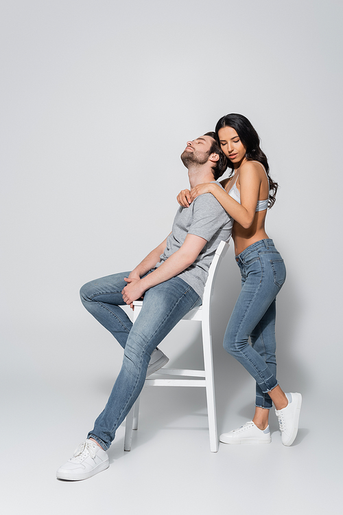 full length view of brunette woman in jeans and bra hugging man sitting on chair on grey