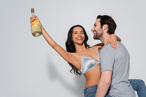 happy man in t-shirt holding sexy woman with champagne bottle on grey