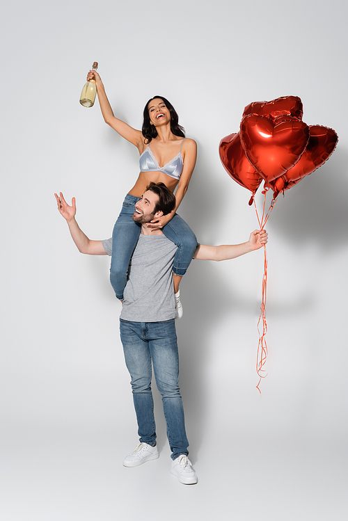 happy man with red balloons piggybacking woman in bra holding champagne bottle on grey