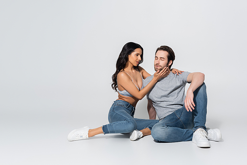 full length view of passionate woman in jeans and bra touching face of young man while sitting on grey