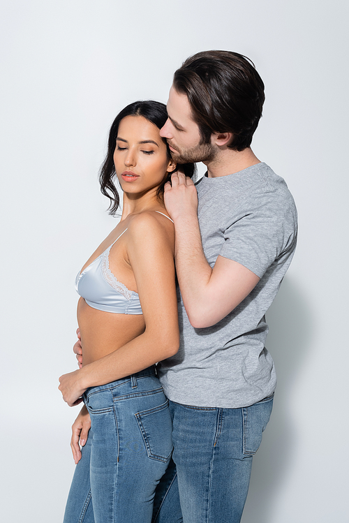 man in t-shirt hugging seductive woman in bra and touching her hair on grey