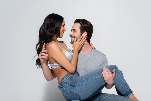 happy man in t-shirt holding woman in jeans and bra touching his face on grey