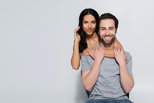 joyful man in t-shirt smiling at camera near seductive woman embracing him on grey