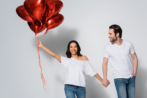 cheerful woman with heart-shaped balloons holding hands with young boyfriend on grey