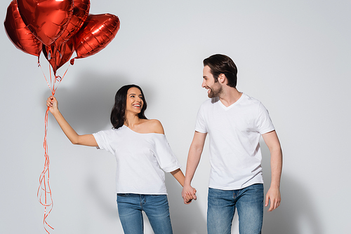 happy young couple in white t-shirts holding hands and looking at each other near heart-shaped balloons on grey