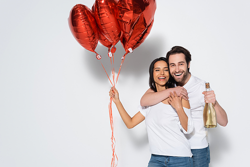 happy man with champagne bottle embracing woman with red heart-shaped balloons on grey