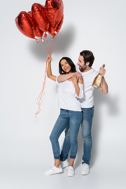 happy man with champagne bottle embracing girlfriend with heart-shaped balloons on grey