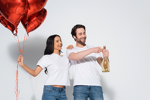 smiling man opening champagne bottle near happy woman with red balloons on grey