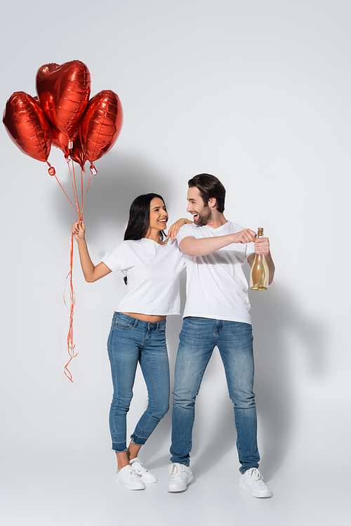 cheerful man opening champagne bottle near girlfriend with heart-shaped balloons on grey