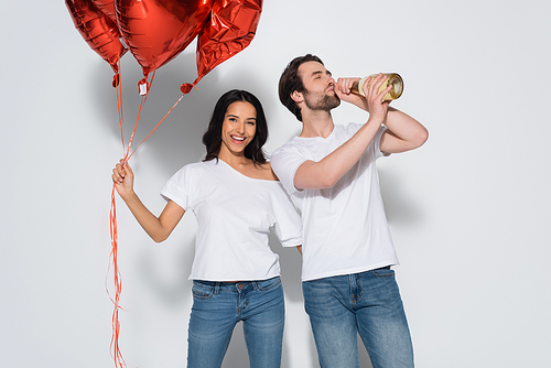 cheerful woman with red balloons  near man drinking champagne from bottle on grey