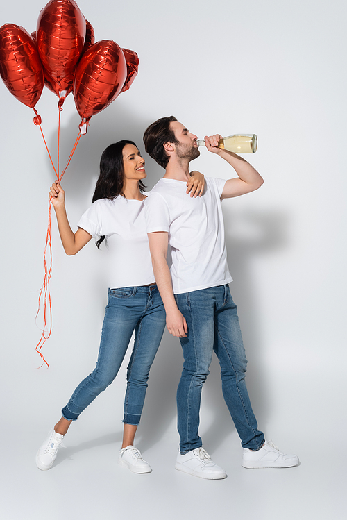 full length view of joyful woman with red heart-shaped balloons near man drinking champagne from bottle on grey