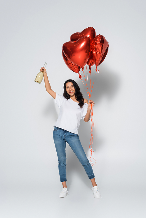 full length view of happy and trendy woman with red heart-shaped balloons and champagne bottle on grey
