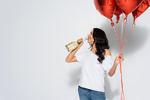 pretty brunette woman with closed eyes holding red balloons while drinking champagne from bottle on grey