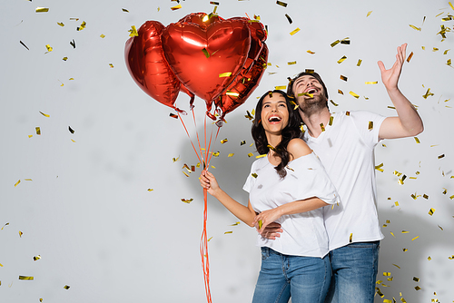 excited couple with heart-shaped balloons laughing under falling confetti on grey