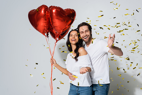 happy woman with red heart-shaped balloons smiling near boyfriend and confetti on grey