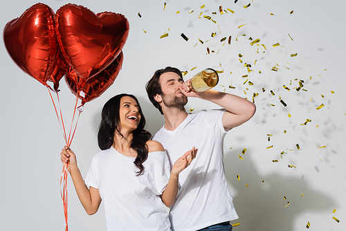 excited woman holding heart-shaped balloons and man drinking champagne from bottle near confetti on grey