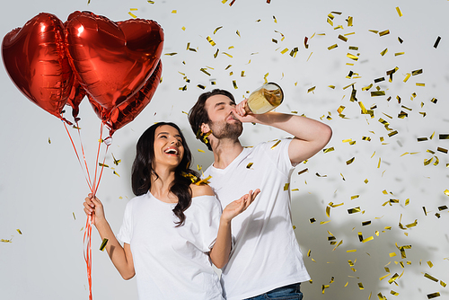 man drinking champagne under falling confetti near excited woman with heart-shaped balloons on grey