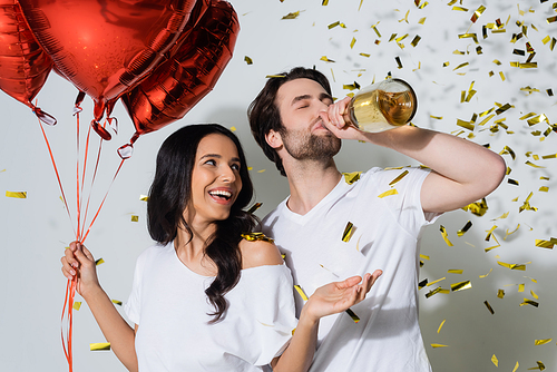 cheerful woman holding red balloons near boyfriend drinking champagne from bottle on grey