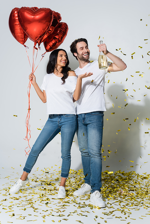 happy woman with heart-shaped balloons pointing at champagne bottle in hand of boyfriend on grey